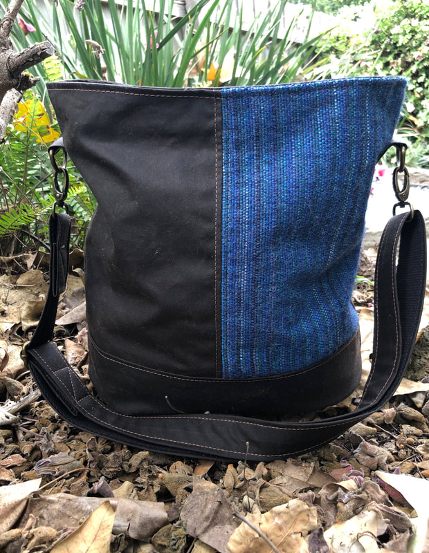 Bag sitting in a garden. Bag has a blue toned woven panel on the right hand side and dark brown canvas panel on the left hand side. Link Browse Bags, Wallets & Accessories
