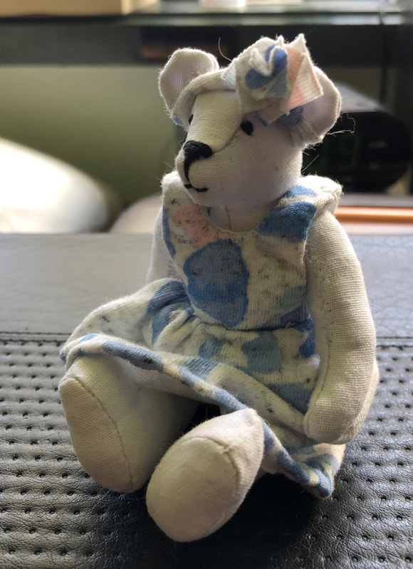 Mini bear (approx 9cm tall) made from white cotton fabric. Bear is wearing a blue, pink & white patterned dress and has a small flower headband